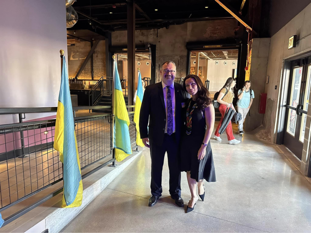 Two people smiling with Ukraine flags behind them.